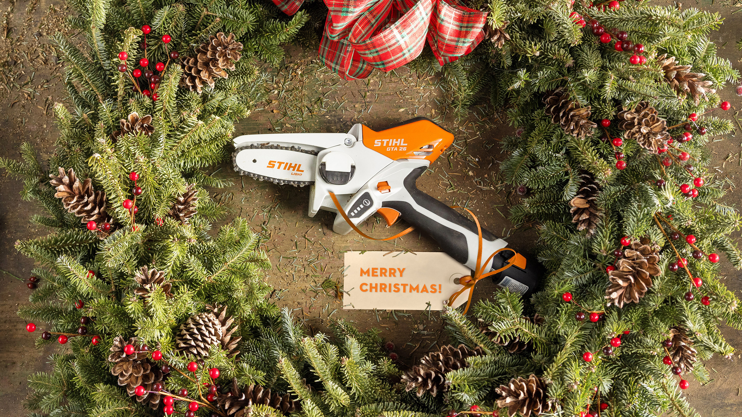 STIHL GTA 26 într-o coroniță de Advent, înconjurat de verdeața bradului, conuri de brad și o panglică cu o felicitare de Crăciun
