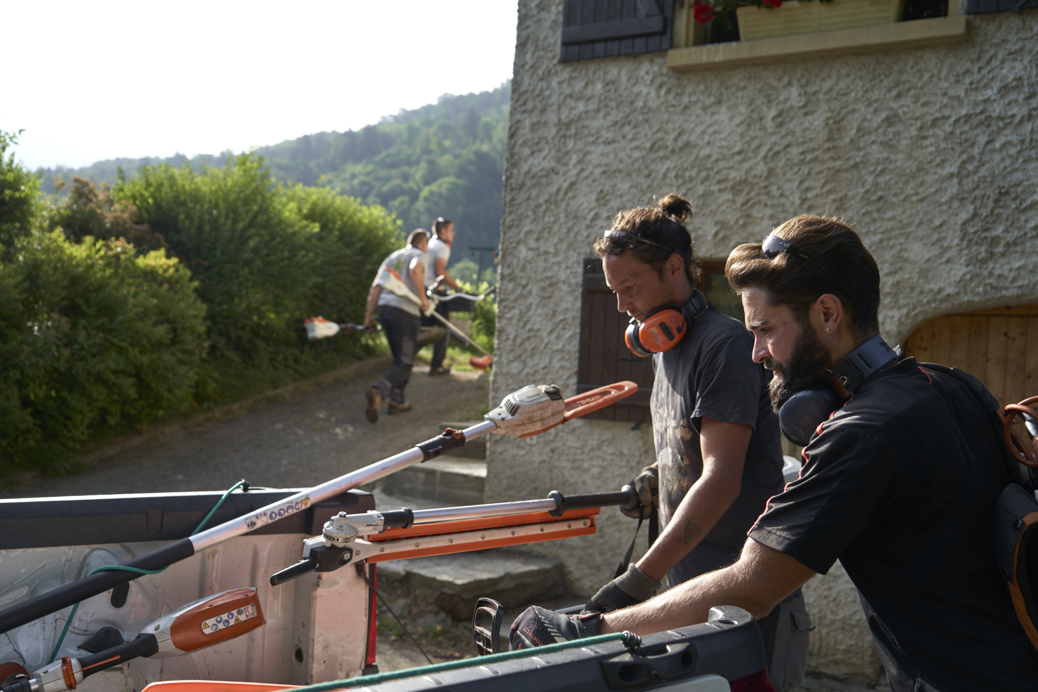 Doi peisagiști descarcă utilajele cu acumulator STIHL dintr-o mașină, cu alți lucrători în fundal. 