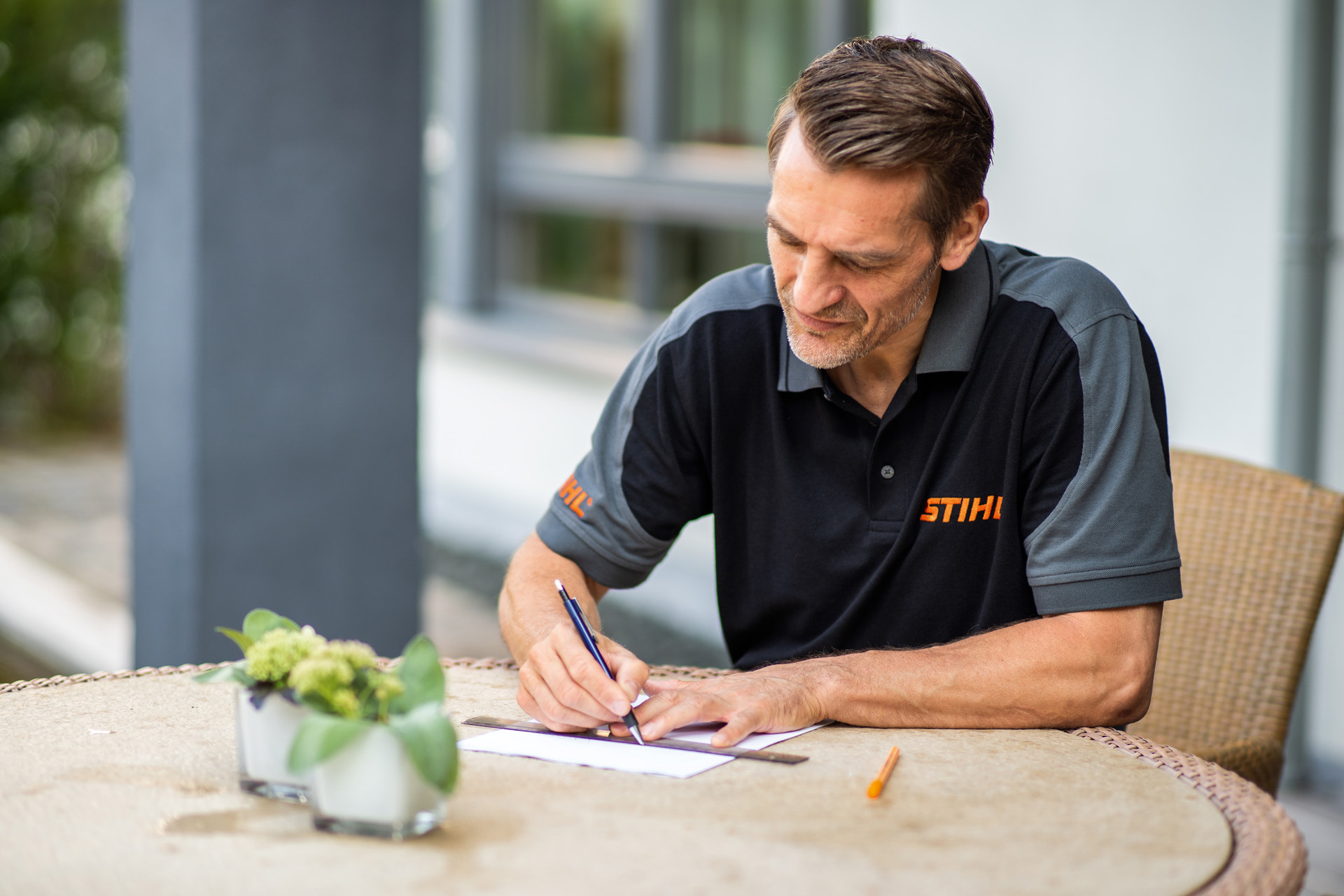 Distribuitorul STIHL stă așezat la masă în grădină și creează planul pentru suprafața de gazon care urmează să fie tunsă de către robotul de tuns iarba iMOW
