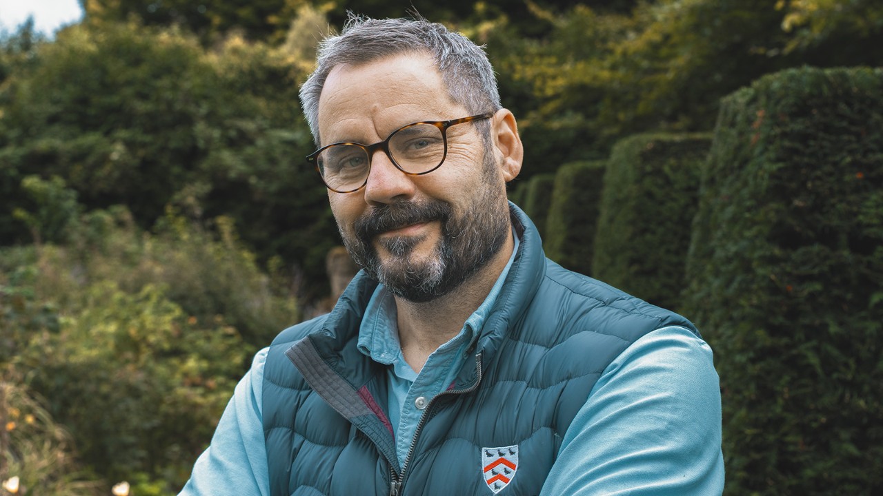 Portret al șefului Grădinilor și Terenurilor de la Worcester College, în picioare, în aer liber, într-o grădină amenajată, purtând ochelari și ținută profesională de lucru.