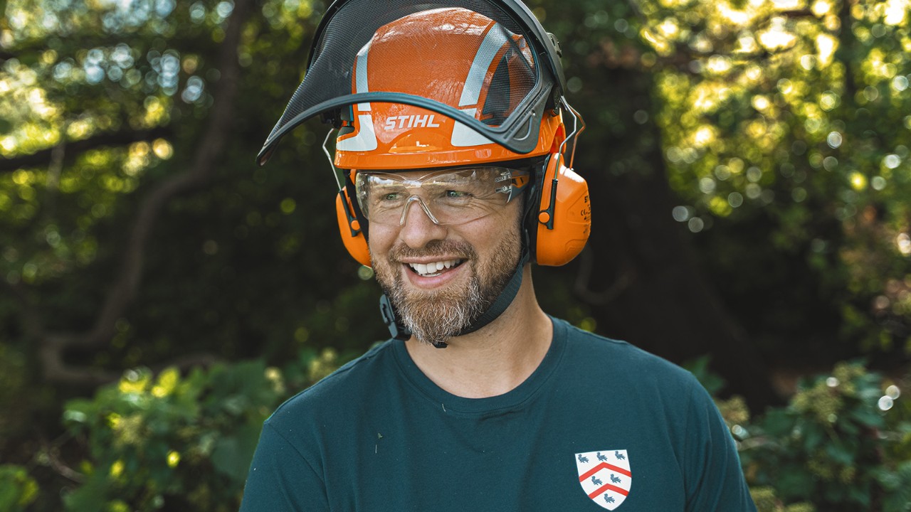Portretul unui arborist de la Colegiul Worcester, purtând cască de protecție și îmbrăcăminte de lucru, stând în aer liber într-un cadru împădurit.