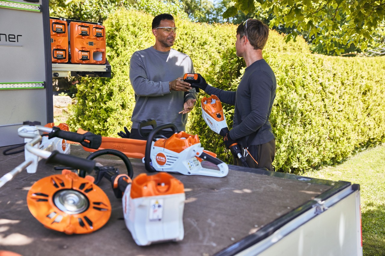 Un bărbat înmânează un acumulator STIHL colegului său, cei doi se află în spatele zonei de încărcare a unui vehicul, pe aceasta se află, printre altele, bottTainer și un motoferăstrău cu acumulator STIHL, precum și alte utilaje