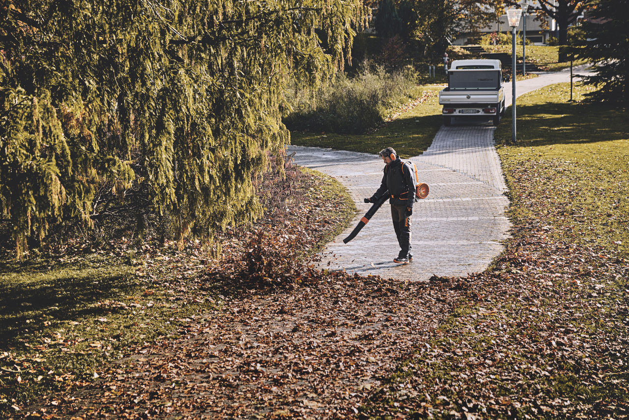 Un bărbat în echipament de protecție îndepărtează frunzele de pe o alee din parc folosind o suflantă de frunze cu acumulator