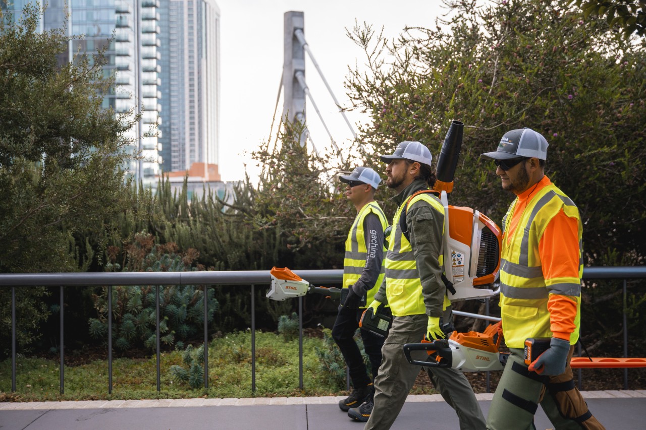 Trei lucrători LaGa merg printr-un oraș purtând îmbrăcăminte de lucru și cărând utilaje STIHL.