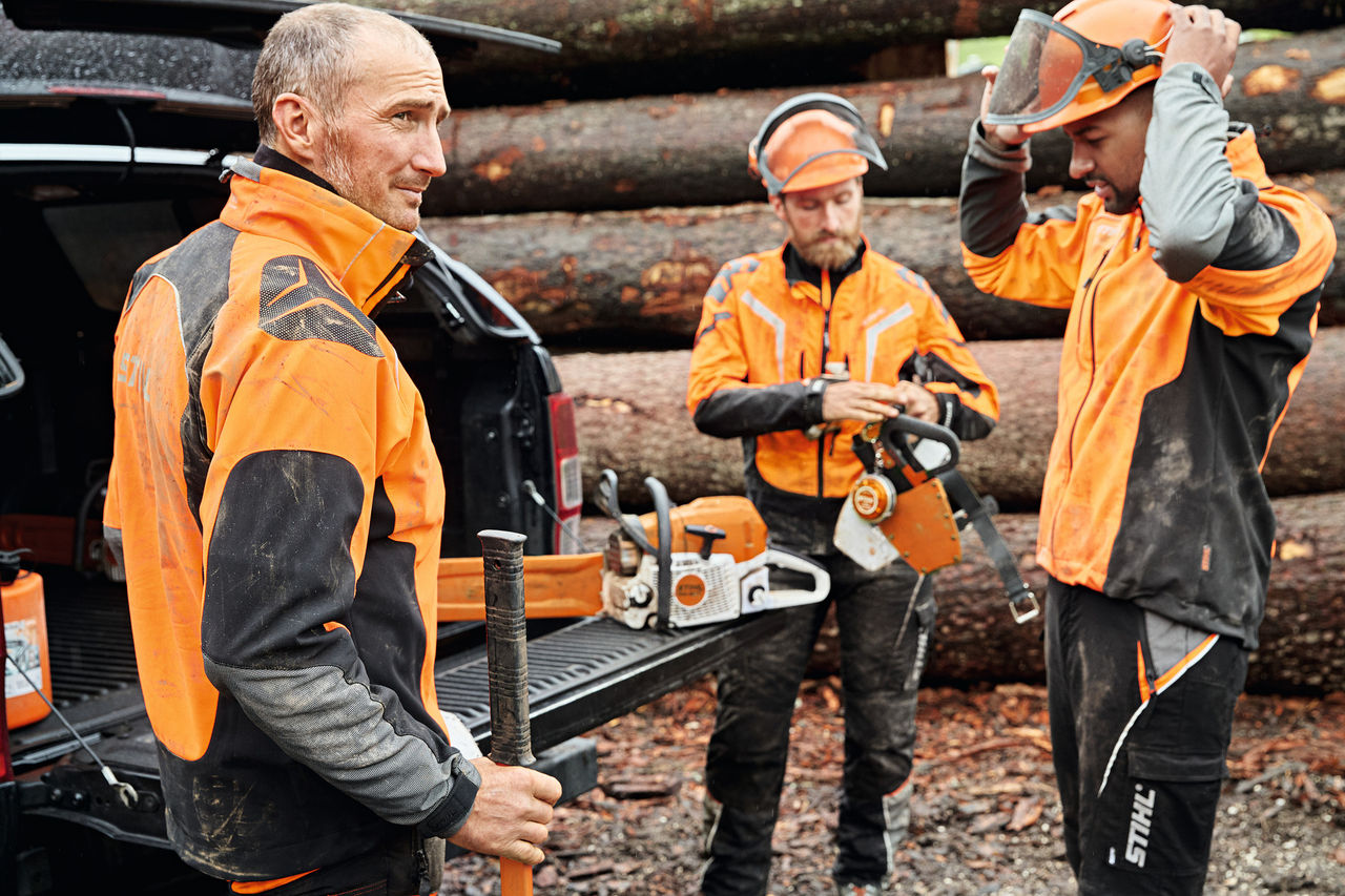 Trei bărbați îmbrăcați în echipament de protecție STIHL cu utilaje cu acumulator STIHL în fața zonei de încărcare a unei mașini
