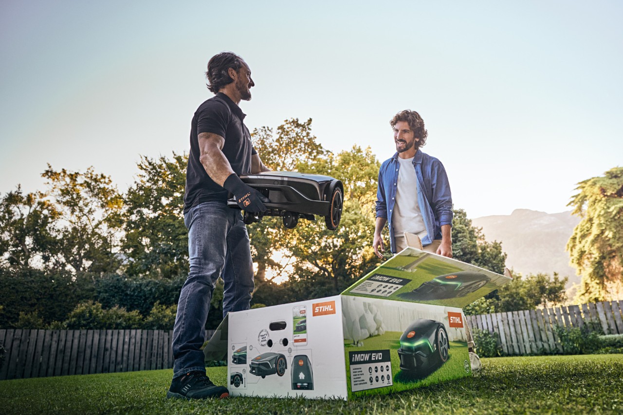 Un distribuitor STIHL se află în grădina unui client cu un iMOW® EVO în mână și o cutie iMOW® în fața sa și dorește să asiste la instalare