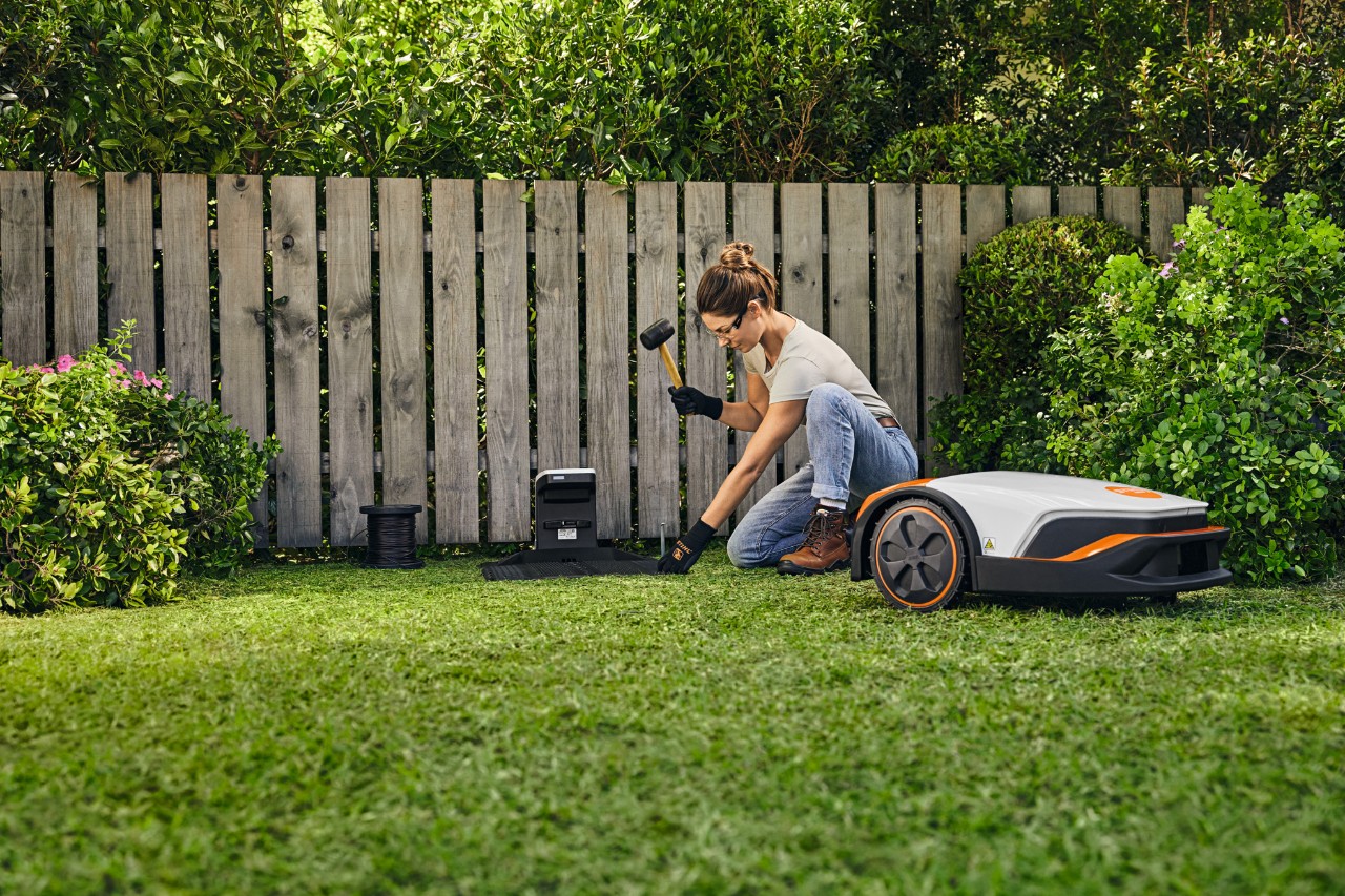 O femeie instalează pe cont propriu stația de andocare pentru robotul STIHL iMOW® în fața unui gard din grădina sa.